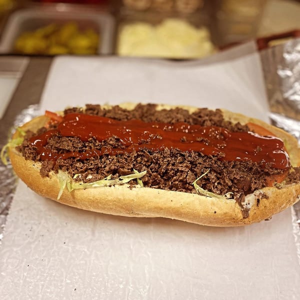 A close-up of a cheesesteak sandwich from Robola Pizzeria, topped with ketchup, lettuce, and tomatoes.