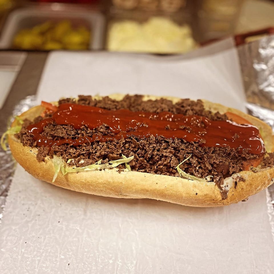A close-up of a cheesesteak sandwich from Robola Pizzeria, topped with ketchup, lettuce, and tomatoes.